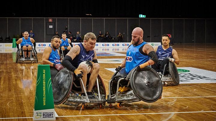 Pictured are a man and woman playing wheelchair rugby in a stadium. Behind them are several other players in wheelchairs